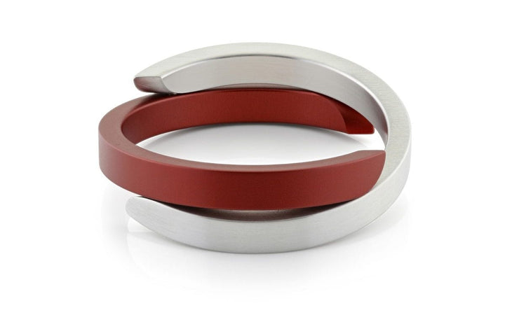 De Armband Cs gerelateerd Rood A1R van CLIC Creations is een strakke, moderne armband met verweven rode en zilveren banden, die een uniek tweekleurig cirkelvormig ontwerp laten zien. Handgemaakt in Nederland, de banden overlappen aan de uiteinden op een reflecterend wit oppervlak.