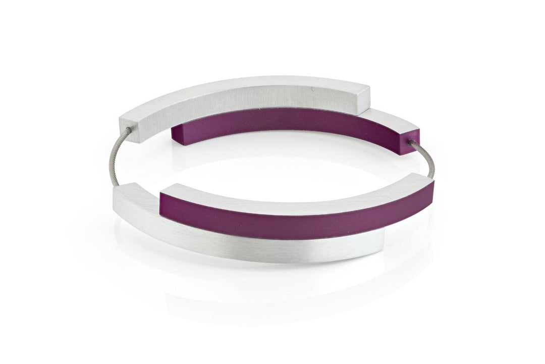 De Armband Vier bogen Paars A32P van CLIC Creations is een moderne armband gemaakt in Nederland. Het heeft twee gebogen zilveren metalen bogen en twee paarse bogen verbonden door een dunne, flexibele draad met een magnetische sluiting, die een cirkelvormig ontwerp vormen op een witte achtergrond.