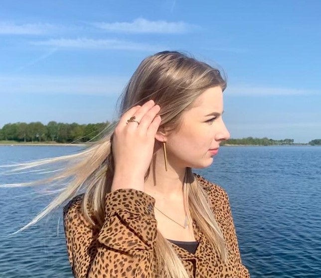 Een persoon met lang blond haar staat bij het water en raakt zijn haar aan, versierd met Oorhanger lange staafjes O38 van CLIC Creations. Ze dragen een shirt met luipaardprint, kijken zijwaarts onder een helderblauwe lucht, met groen zichtbaar in de verte.