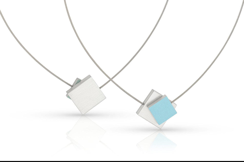 Collier Vierkant met gekleurd vierkant C263B van CLIC Creations bestaat uit twee minimalistische kettingen: één met een blauw vierkant over een wit vierkant, en een andere met een effen wit vierkant. Beide hangers hangen aan dunne zilveren koorden met magnetische sluitingen, prachtig weerspiegeld op een wit oppervlak.