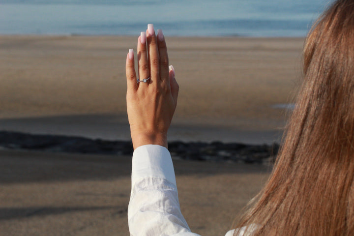 De hand van een vrouw versierd met de See You Isabelle | AsRing Zilver is opgeheven, tegen een achtergrond van een zandstrand en de oceaan. Ze draagt een wit shirt met lange mouwen en haar lange bruine haar is gedeeltelijk zichtbaar.