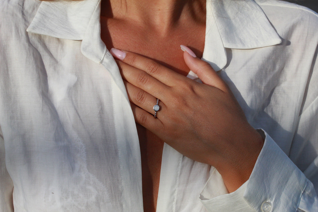 Een persoon met verzorgde nagels, in een wit licht open shirt, raakt zijn borst aan en toont elegant de See You Isabelle | AsRing Zilver. Deze sterling zilveren ring is versierd met een ronde witte steen, die zijn look perfect aanvult.