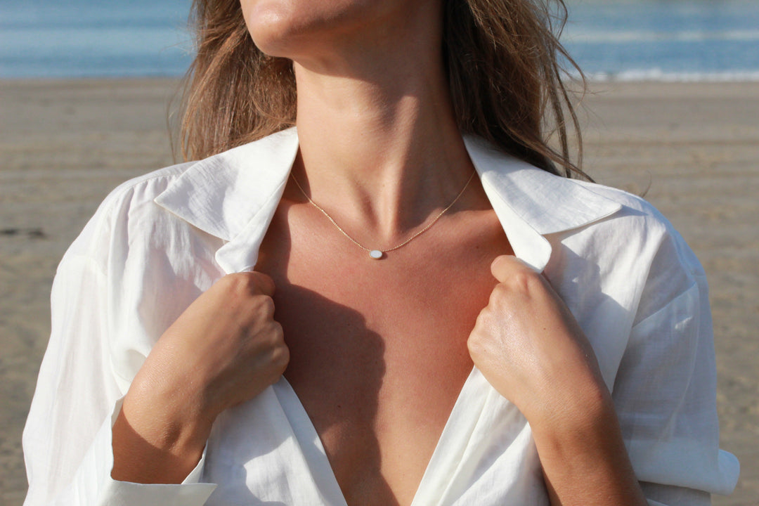 Een persoon in een wit shirt staat op het strand en houdt voorzichtig de kraag open. Ze dragen de Luna | AsKetting Zilver, een minimalistische klassieke ovale ketting van See You, tegen een serene achtergrond van oceaan en zand.