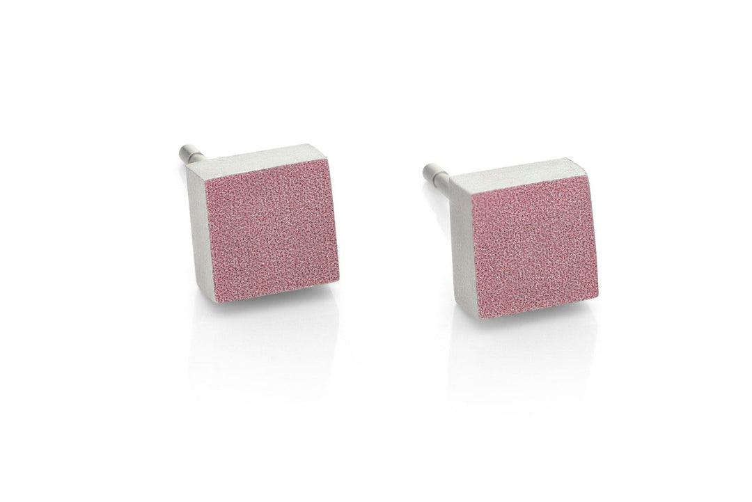 Close-up van CLIC Creations Oorsteker Klein vierkant Roze O37RO, handgemaakt in Nederland. Deze hypoallergene oorbellen hebben een getextureerd matroze oppervlak en metalen staafjes. Naast elkaar geplaatst op een witte achtergrond, weerspiegelen ze subtiel eronder.