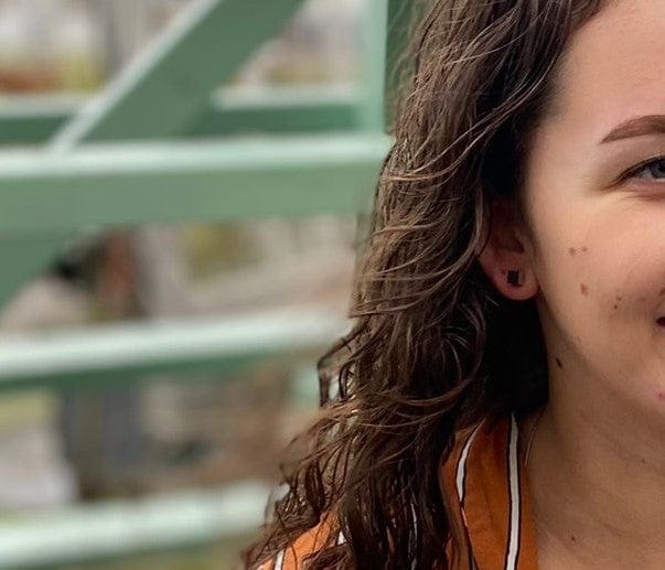 Close-up van een persoon met golvend bruin haar die CLIC Creations Oorsieraad Kubusjes Oranje O28O en een oranje-wit shirt draagt, van rechts voor een groen metalen bouwwerk buiten. Slechts de helft van het gezicht en de schouder zijn zichtbaar.