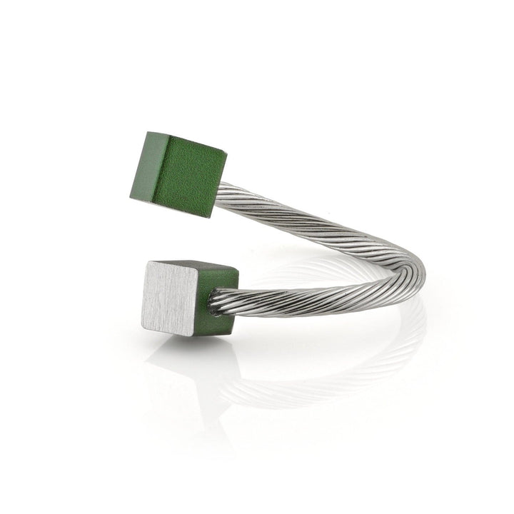De Ring Kubusjes Groen R4GR van CLIC Creations is een gedraaide zilveren kabelarmband met open uiteinden, versierd met een matte groene kubus en een glanzende zilveren kubus. Het rust op een reflecterend wit oppervlak, wat het unieke ontwerp benadrukt.