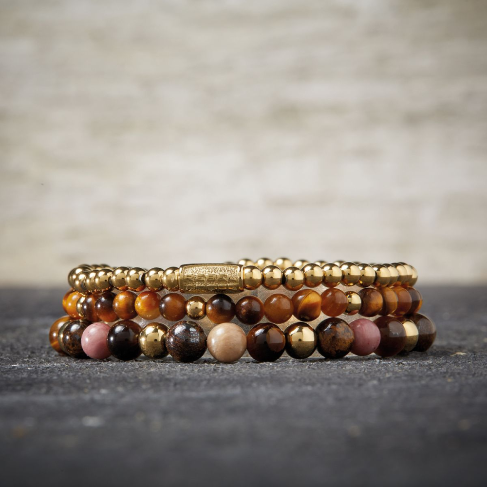 Vier Rebel & Rose "Tiger Lily" armbanden voor dames, met 4 mm kralen en 18 kt geelgouden ion-plating, zijn stijlvol gestapeld op een donker gestructureerd oppervlak met een vage lichte achtergrond.