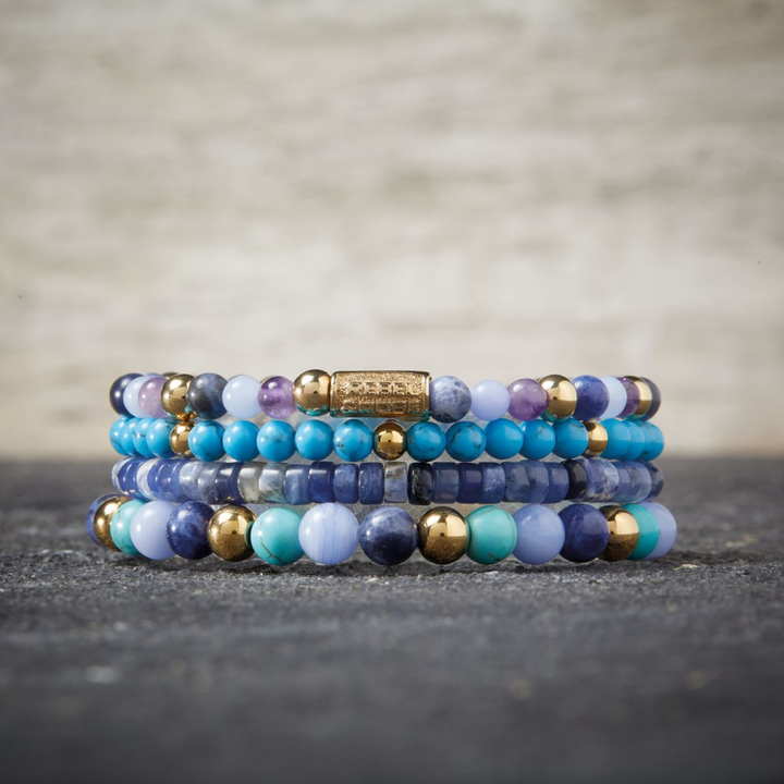 Een stapel kralenarmbanden met de Rebel & Rose Winter Blues - 18 kt geelgoud geïoniseerd 4mm Kralen Armband Dames wordt weergegeven op een grijs oppervlak met textuur en een vage lichte achtergrond.
