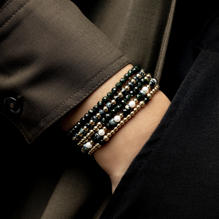 Een close-up van een pols die de Rebel & Rose Green Glam Rocks Gold 4mm Kralen Armband Dames draagt, met groene zandsteen kralen met zwarte, gouden en witte parelachtige accenten, gestyled met een donkere getailleerde outfit.