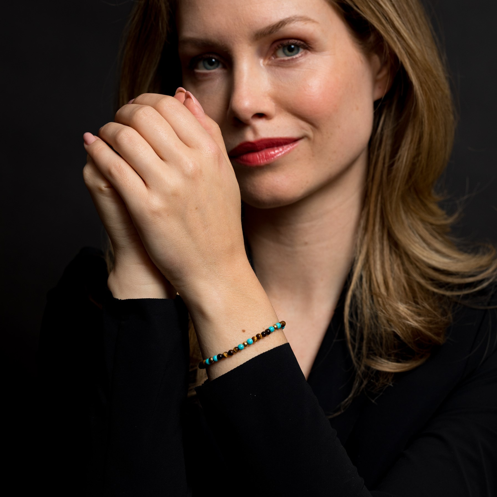 Een vrouw met lichtbruin haar in een zwart topje poseert tegen een donkere achtergrond, glimlacht zachtjes terwijl ze de Rebel & Rose Mix Tijger Turkoois 18 kt geel goud geïoniseerd 4mm kralen armband dames laat zien - een elegante toevoeging aan haar collectie.