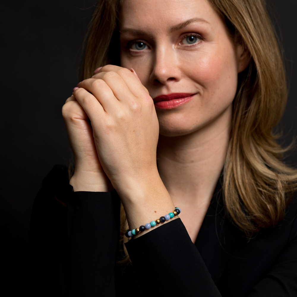 Een blonde vrouw in een zwart topje glimlacht zachtjes, terwijl ze haar Rebel & Rose Winter Blues - 6mm Kralen Armband Dames met turquoise agaat sodaliet stenen om haar pols laat zien tegen een donkere achtergrond.