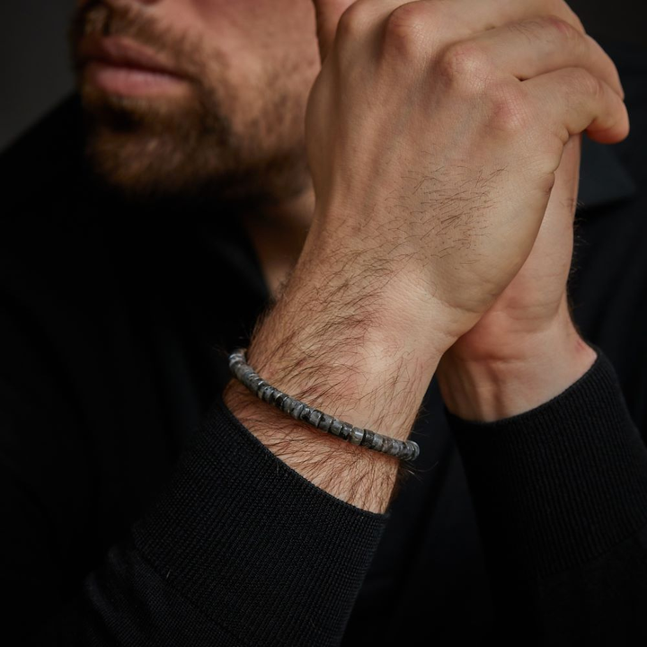 Een man in een donker shirt met lange mouwen omklemt zijn handen en toont de Rebel & Rose Slices - Grey Seduction 6mm heren kralen armband om zijn pols. Zijn gezicht is gedeeltelijk zichtbaar met een getrimde baard.