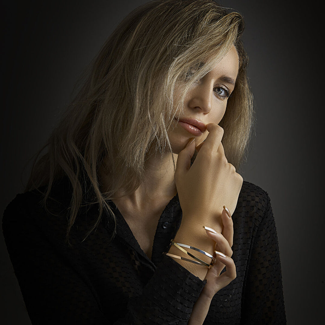 Een vrouw met golvend blond haar in een zwart topje met structuur kijkt naar de camera. Ze laat haar hand op haar kin rusten en draagt de armband scharnier massief vlakke buis 3 x 61 mm Zilgold van Van der Kooij Collectie om haar pols, tegen een donkere achtergrond.