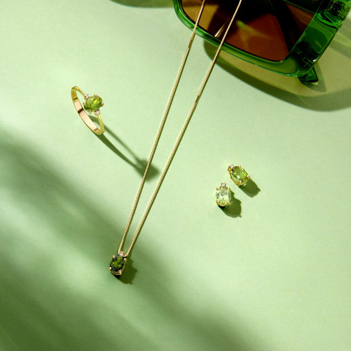 Van der Kooij Collectie’s 14K geelgouden collier toermalijn en diamant (0.02ct H SI, 40-42-44 cm) schittert met feitelijke ring, ketting en oorbellen op een lichtgroen oppervlak naast een transparante groene zonnebril.