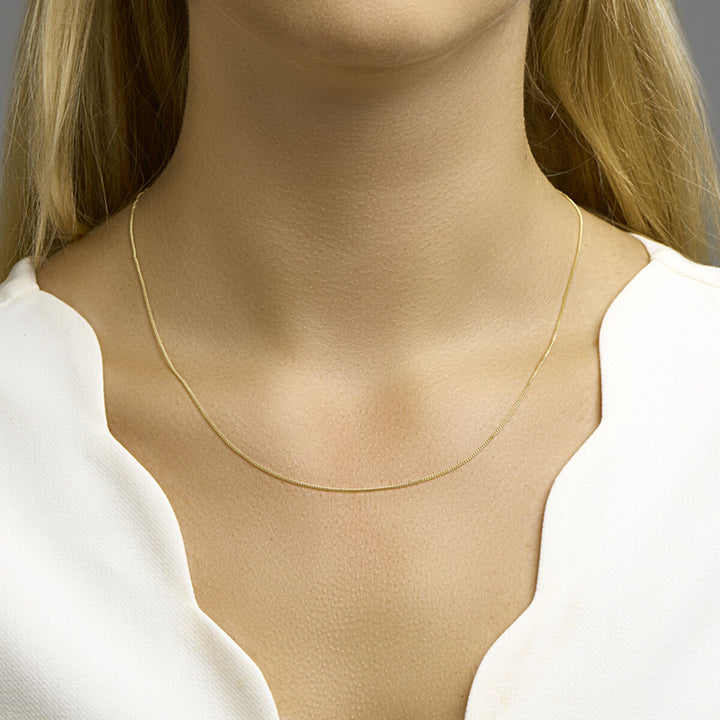 Een vrouw met lang blond haar draagt het Van der Kooij Collectie collier gourmette 1,0 mm 14K geelgoud en een wit topje met schulprand, vanaf de schouders omhoog afgebeeld met het collier rustend op haar sleutelbeen.