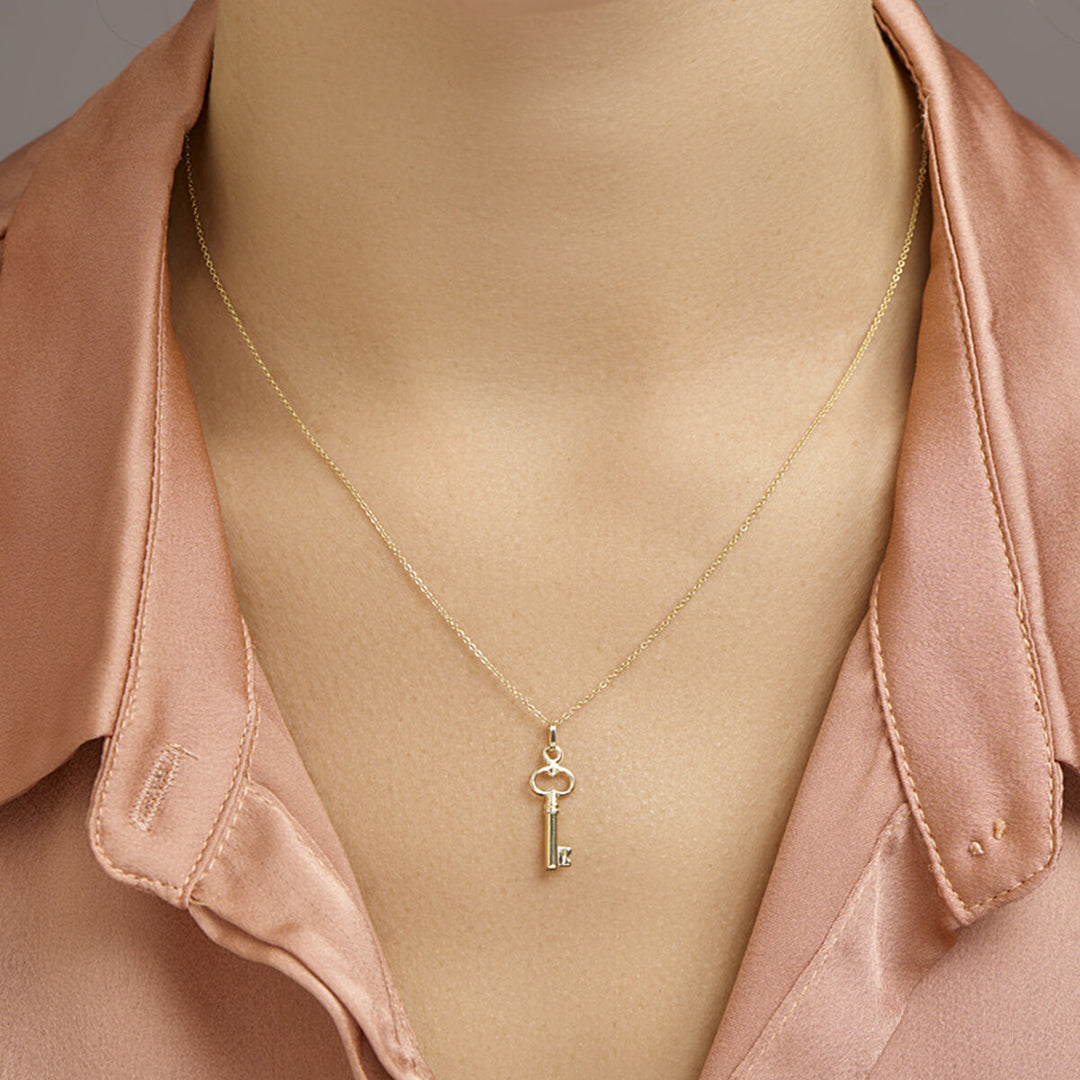 Een persoon in een lichtroze overhemd toont de Van der Kooij Collectie hanger sleutel 14K geelgoud-een delicate sleutelvormige geelgouden hanger-rustend op het midden van zijn borst.