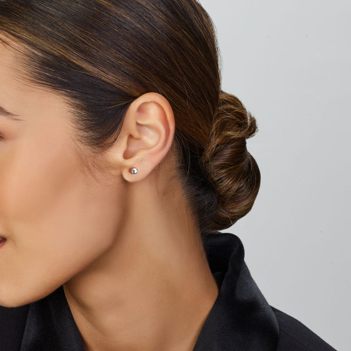 Een vrouw met donker haar in een lage knot draagt de oorknoppen bolletje zilver gerhodineerd van Van der Kooij Collectie, gecombineerd met een zwart topje. Het close-up zijaanzicht speelt zich af tegen een lichtgrijze achtergrond.