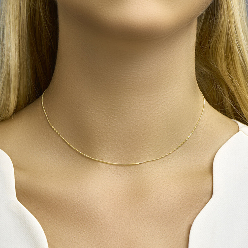 Een vrouw met blond haar draagt het Van der Kooij Collectie collier gourmette 1,0 mm 14K geelgoud om haar hals. Ze draagt een wit topje met V-hals tegen een neutrale achtergrond.