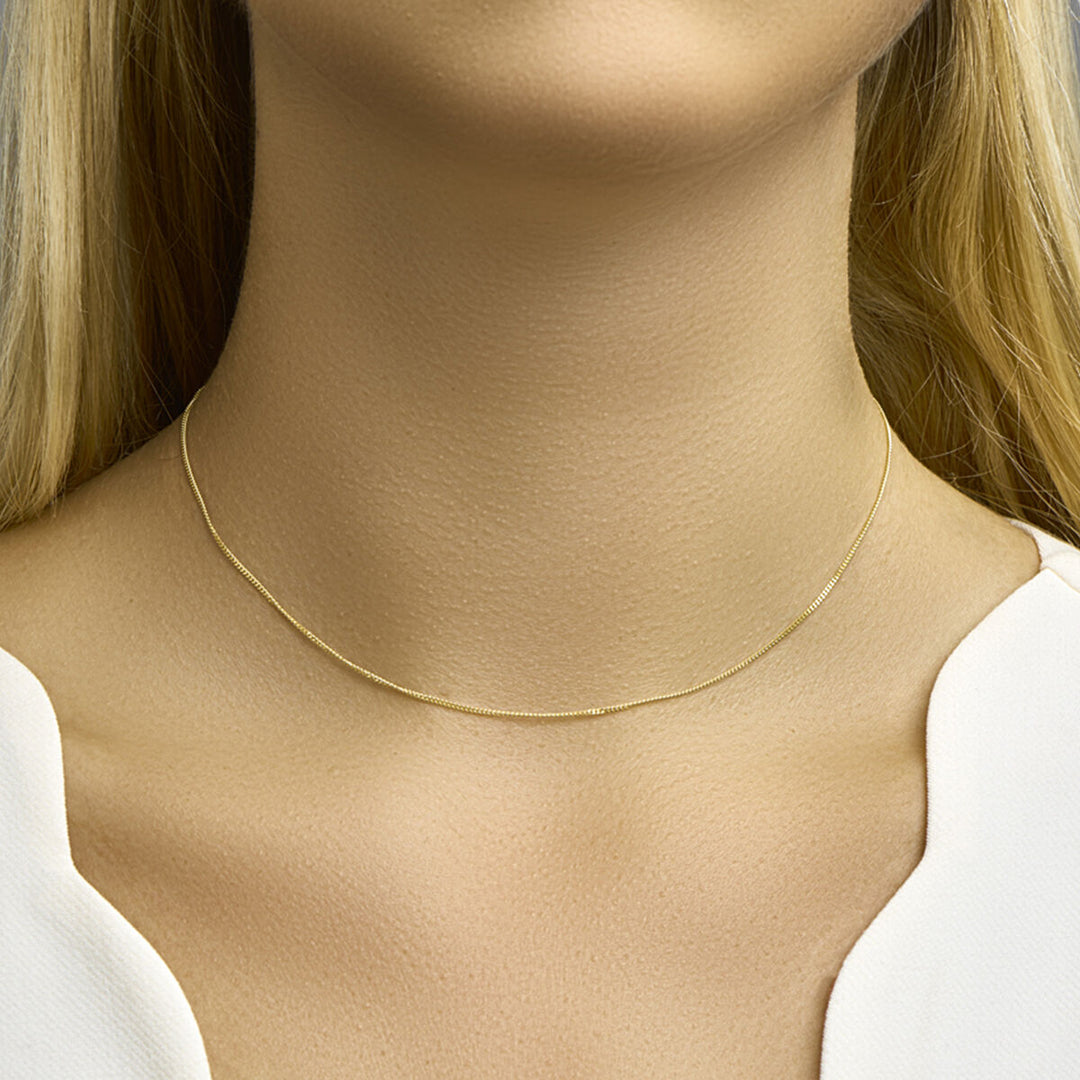 Een vrouw met blond haar draagt het Van der Kooij Collectie collier gourmette 1,0 mm 14K geelgoud om haar hals. Ze draagt een wit topje met V-hals tegen een neutrale achtergrond.