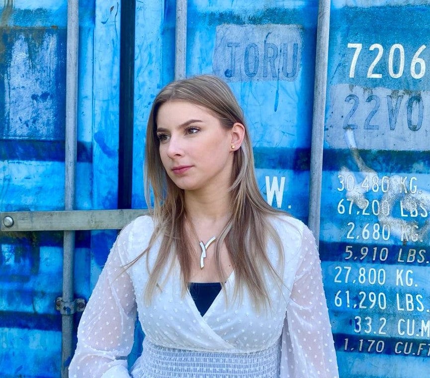 Een vrouw met lang blond haar staat voor een blauwe scheepscontainer versierd met nummers en tekst. Ze draagt een witte blouse met stippen over een zwart topje en draagt een zilveren ketting met CLIC Creations Collier Bogen op elkaar Goud|Geel C254G met een magnetische sluiting, terwijl ze naar de zijkant kijkt.