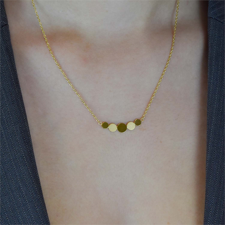 Een close-up van iemand die de Boccia Titanium Figura Collier gp 08031-02 draagt, met vijf glanzende, platte ronde schijven in verschillende maten van Boccia. De glimmende hanger contrasteert prachtig met een donkere, getextureerde top of jasje.