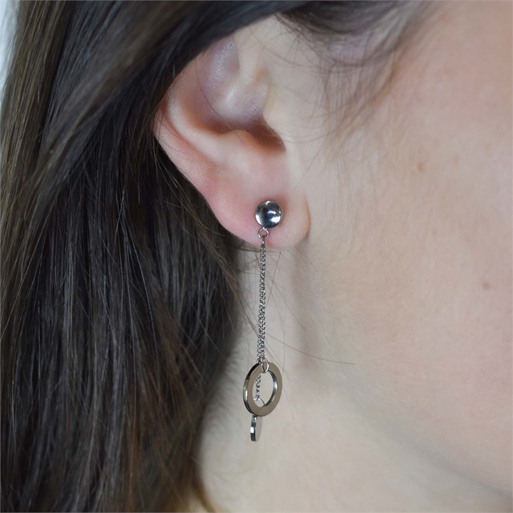 Een close-up van het oor van een persoon met Boccia Titanium Figura Oorstekers 05042-01 van Boccia, met daarop een ronde oorbel, een dunne ketting en een hangende ronde hanger uit de Figura-collectie. Gedeeltelijk zichtbaar haar en onscherpe achtergrond.