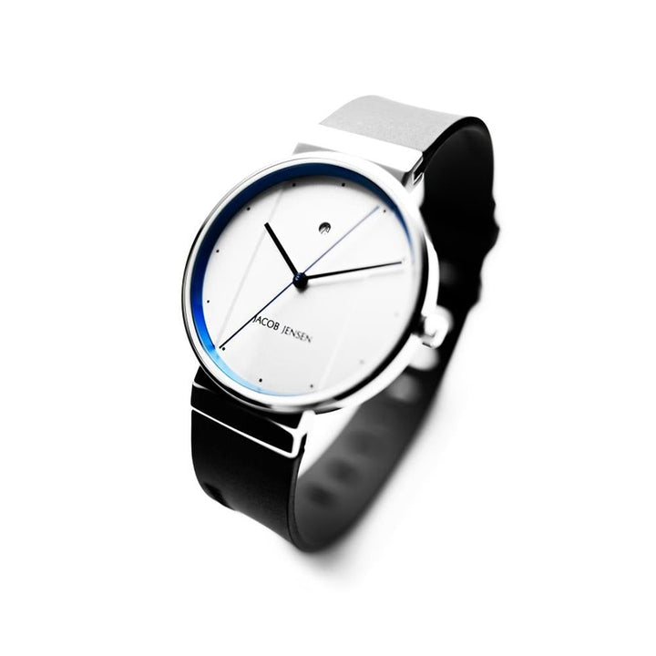 Het Jacob Jensen 750 horloge heeft een modern design met een witte wijzerplaat, minimalistische dunne zwarte wijzers en een quartz uurwerk. Het heeft een roestvrijstalen zilveren kast en zwarte band, met de merknaam elegant weergegeven op de wijzerplaat.