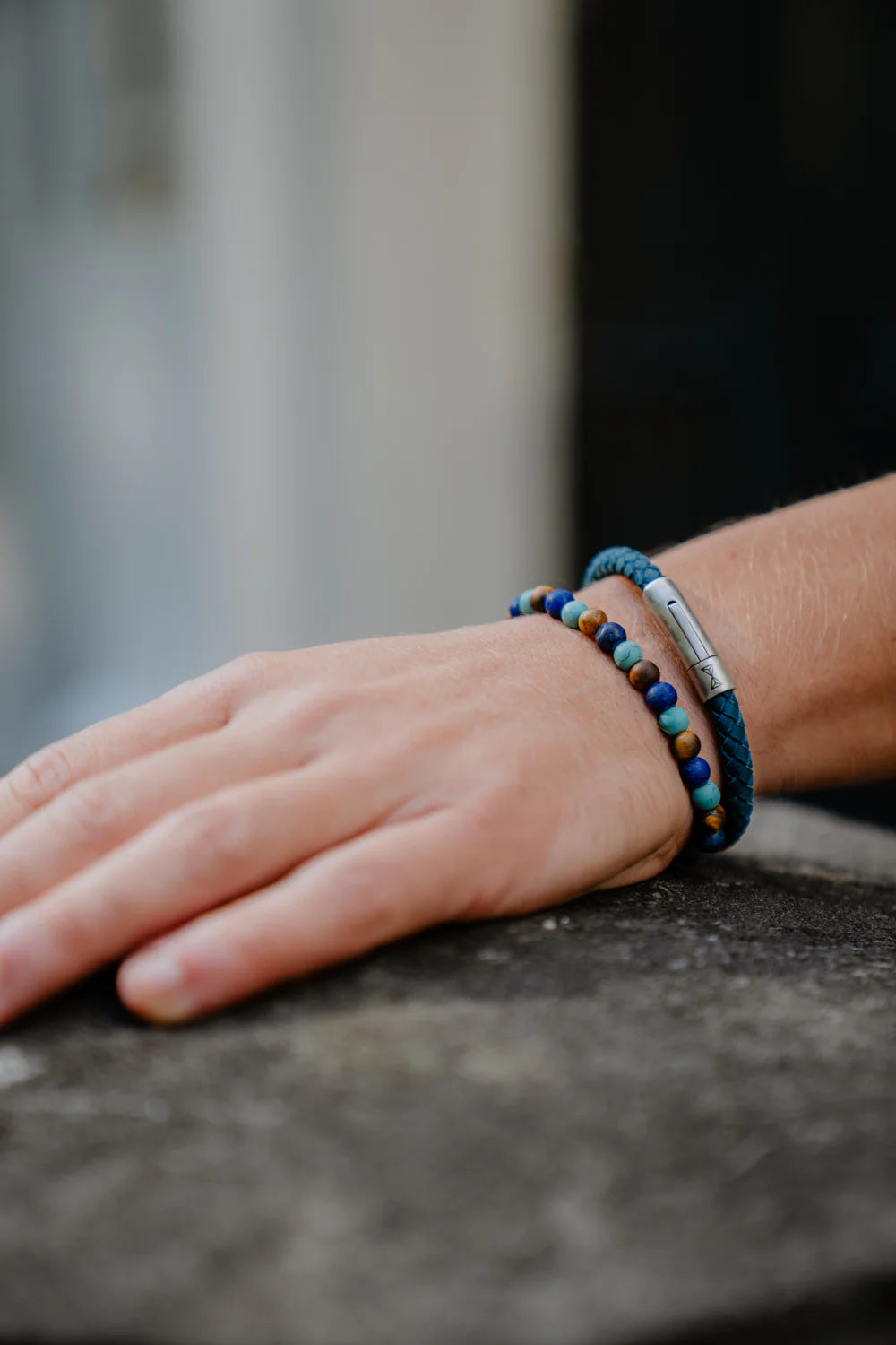 Een hand rust op een steen, versierd met twee armbanden: de Kralen armband heren - Mount Sinai van AZE Jewels, met blauwe en bruine kralen, en een gestructureerde blauwe band met een metalen sluiting die moeiteloze elegantie uitstraalt.