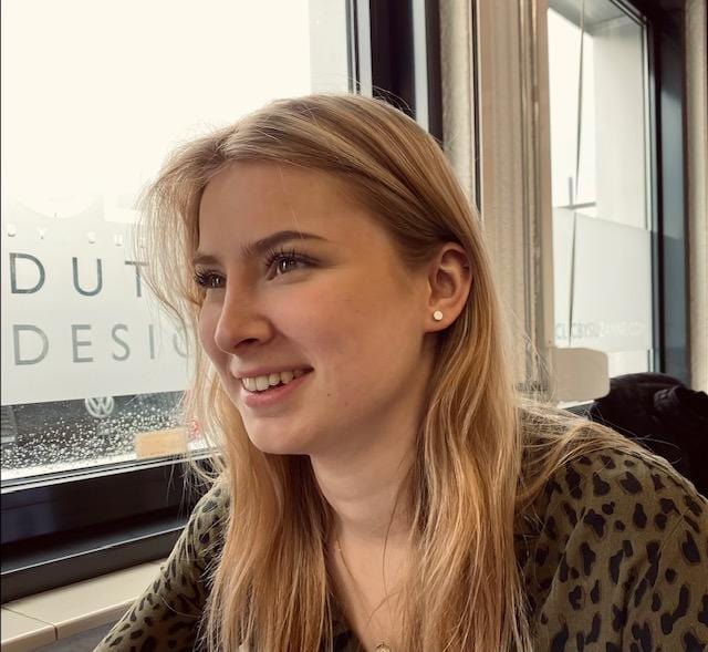 Een persoon met lang blond haar zit bij een raam, glimlachend en zijwaarts starend. Ze dragen een groen gebloemd topje en kleine CLIC Creations Oorsteker Klein rond Roze O36RO oorbellen. Buiten het raam regent het.