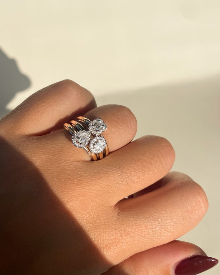 Een hand toont drie 14k gouden ringen, waaronder de opvallende Excellent Jewelry 14k Bicolor Gold Ring met een smaragd geslepen diamant in een entourage, die allemaal schitteren in natuurlijk licht.