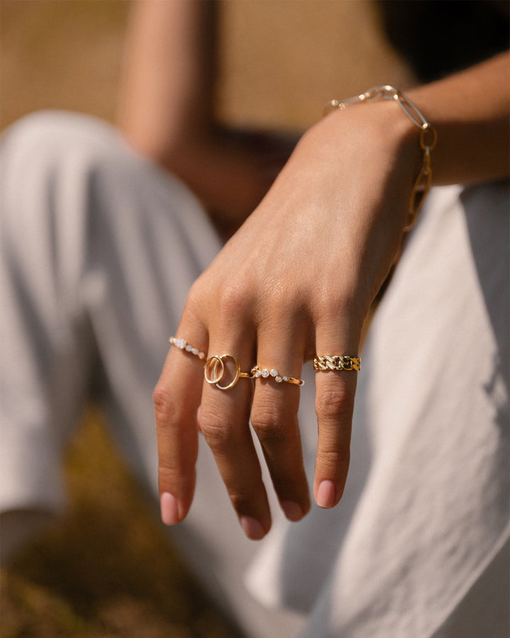 Een close-up van een hand met de Excellent Jewelry 14 Karaat Geelgouden Damesring (6 mm breed, 2,6 gram, eenvoudig ontwerp), rustend op een knie. De persoon draagt een wit broek en heeft kort gemanicuurde nagels; achtergrond is zacht vervaagd.