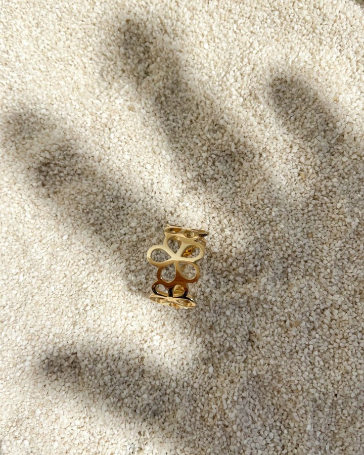 De Geelgouden Damesring met Opengewerkt Bloemenpatroon van Excellent Jewelry, vervaardigd uit 14 karaat goud, ligt op licht zand; haar schaduw versmolten met die van een uitgestrekte hand die ernaar reikt.