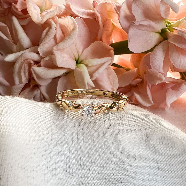 De Elegante Geelgouden Ring met 0,16 krt Briljant Diamant van Excellent Jewelry roest op witte stof, omringd door zachte roze bloemen. Subtiele luxe voor dames straalt uit dit verfijnde sieraad.