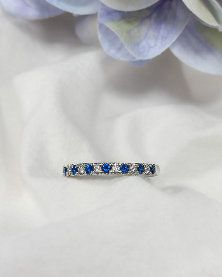 De 14 karaat witgouden rijring met blauwe saffier en 0,14 ct diamanten van Excellent Jewelry schittert op een zacht wit kleed, omgeven door paarse en groene bloemblaadjes in de vervaagde achtergrond.